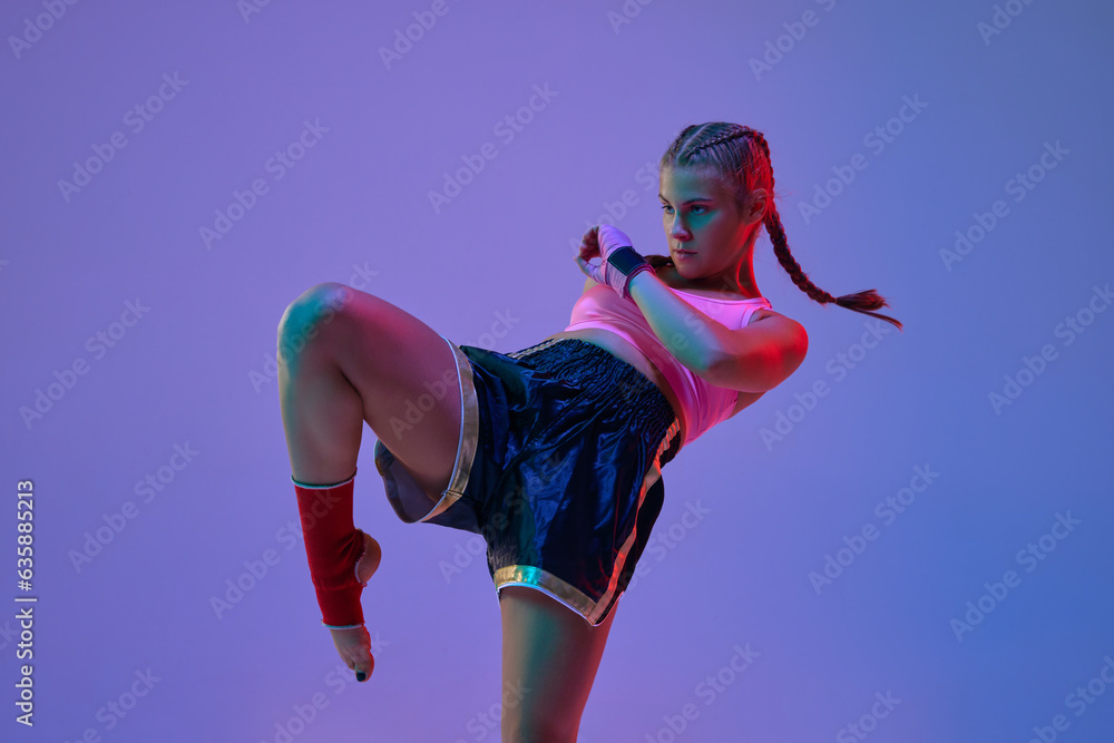 Fototapeta premium Leg kick. Sportive teen girl, mma fighter athlete in motion, training, fighting against purple background in neon lights. Concept of mixed martial arts, sport, hobby, competition, strength, ad
