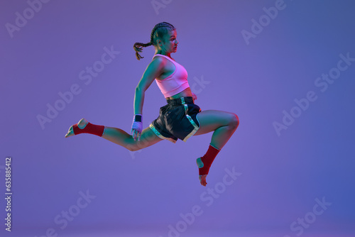 Kick in a jump. Teen girl, mma athlete in motion, training against purple studio background in neon lights. Concept of mixed martial arts, sport, hobby, competition, athleticism, strength, ad