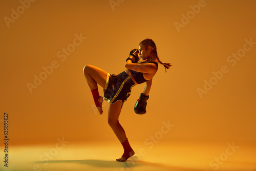 Leg kick. Sportive teen girl, mma fighter athlete in motion, training, fighting against orange studio background in neon lights. Concept of mixed martial arts, sport, hobby, competition, strength, ad photo