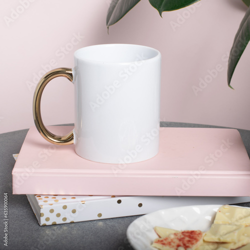 White mug mockup with books and accessories on a table and a ficus plant. White blank cup with golden handle for your design. Empty mockup template