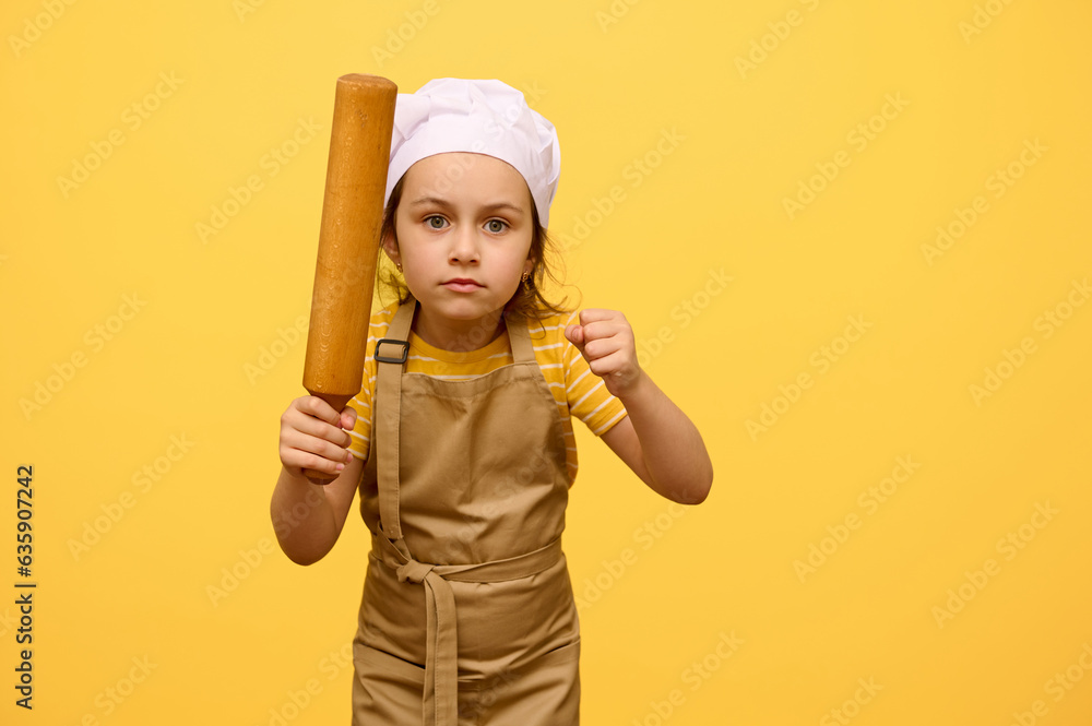 Portrait of a serious angry little child girl in chef's uniform, holding a wooden rolling pin, clenching fists, looking at camera, expressing furiosity, standing isolated over studio yellow background Portrait of a serious angry little child girl in chef's uniform, holding a wooden rolling pin, clenching fists, looking at camera, expressing furiosity, standing isolated over studio yellow background