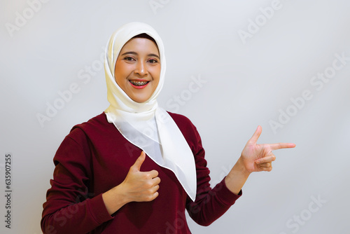 A smiling Asian muslim woman wearing red top and white hijab, pointing to her left, isolated by white background. Indonesia's independence day concept photo