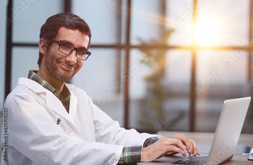 A handsome young doctor sits at a table and smiles friendly while looking at the camera.