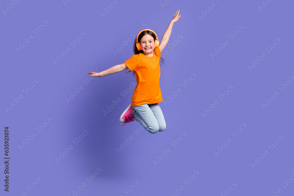 Full length photo of cheerful schoolkid dressed orange t-shirt flying in headphones arms wings isolated on purple color background