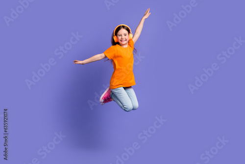 Full length photo of cheerful schoolkid dressed orange t-shirt flying in headphones arms wings isolated on purple color background
