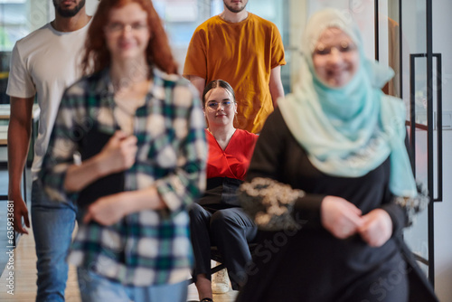 A diverse group of business people walking a corridor in the glass-enclosed office of a modern startup, including a person in a wheelchair and a woman wearing a hijab, showing a dynamic mix of