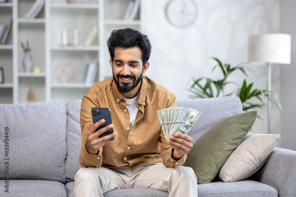 Joyful winner man celebrating victory sitting on sofa at home in living room, hispanic man holding money dollars cash and phone in hands, using smartphone app.