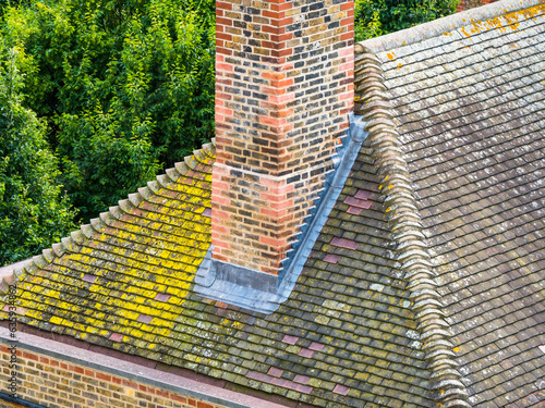 A chimney stack with new lead flashing. Roof tiles aged with moss and yellow lichen. Concept roofing repairs, building maintenance, home improvements, house construction and roof top tile detail.