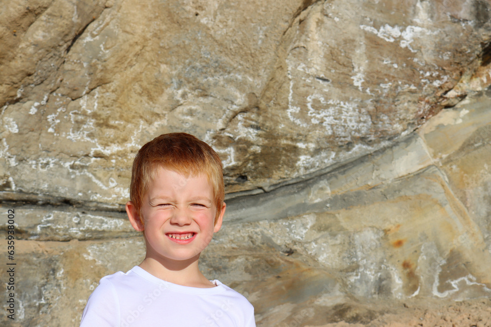 Portrait of a red-haired boy against the background of rocks. Six-year-old boy squints his eyes from the sun
