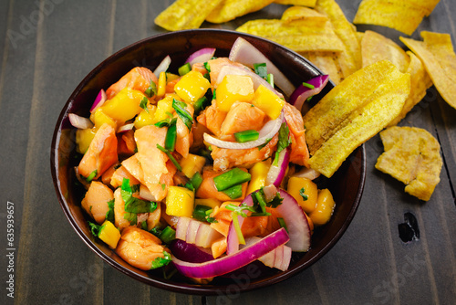 Salmon Ceviche with Mango in a Wooden Bowl with Plantain Chips: A bowl of salmon ceviche surrounded with ingredients on a wooden table