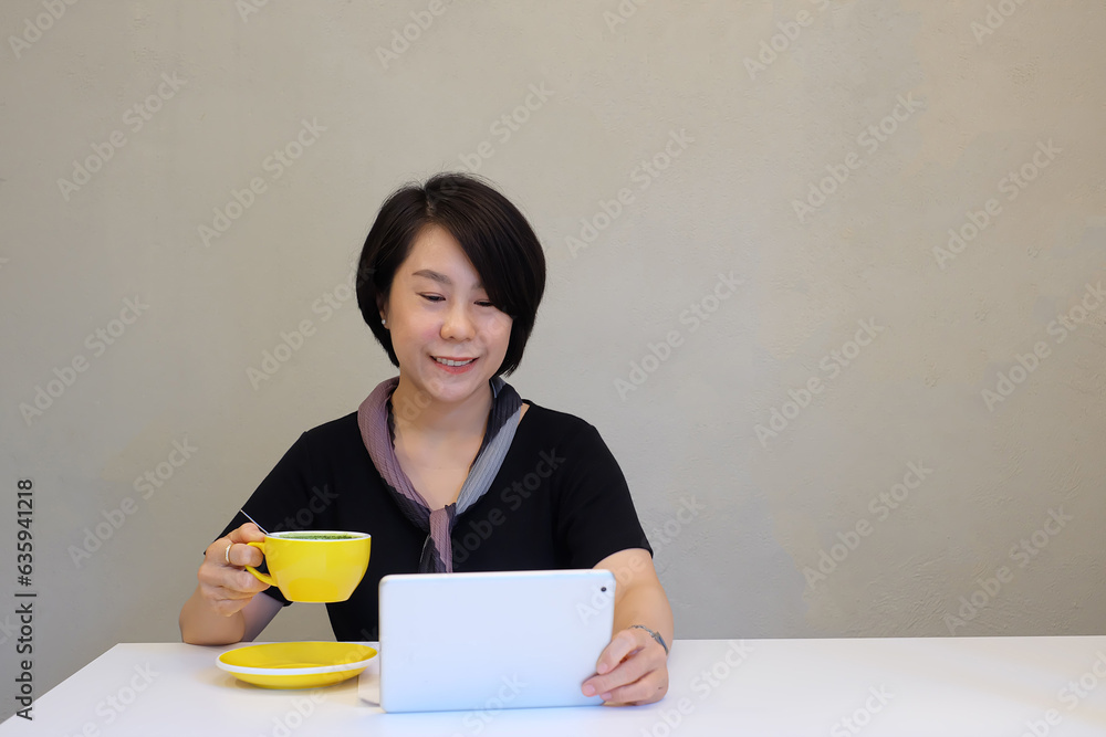 Asian businesswoman or freelancer working on tablet or watching movies, watching news, buying bitcoins, trading stocks, video conference while sipping morning coffee at work desk. Asian businesswoman or freelancer working on tablet or watching movies, watching news, buying bitcoins, trading stocks, video conference while sipping morning coffee at work desk.