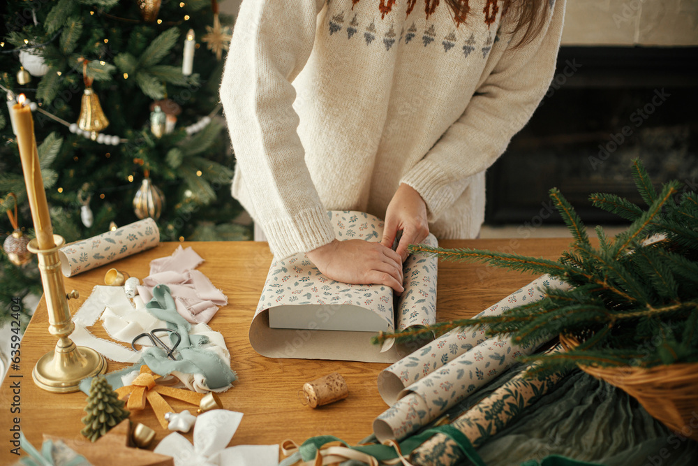 Woman in stylish sweater wrapping christmas gift in paper on wooden table with festive decorations in decorated scandinavian room. Merry Christmas! Hands packing present close up Woman in stylish sweater wrapping christmas gift in paper on wooden table with festive decorations in decorated scandinavian room. Merry Christmas! Hands packing present close up