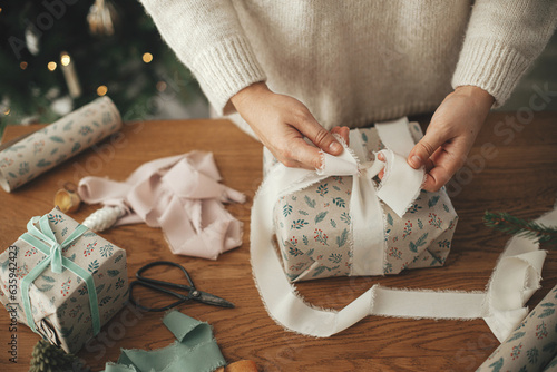 Woman wrapping christmas gift with stylish ribbon on wooden table with festive decorations in decorated scandinavian room. Merry Christmas! Hands packing present close up photo