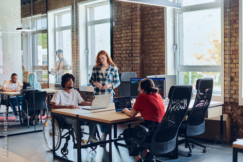 A young business group, including an African American businessman in a wheelchair, collaborates within a modern glass office, actively engaged around a computer and laptop, collectively solving