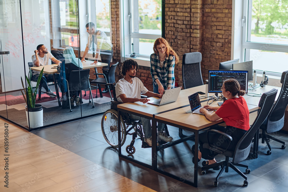 A young business group, including an African American businessman in a wheelchair, collaborates within a modern glass office, actively engaged around a computer and laptop, collectively solving A young business group, including an African American businessman in a wheelchair, collaborates within a modern glass office, actively engaged around a computer and laptop, collectively solving