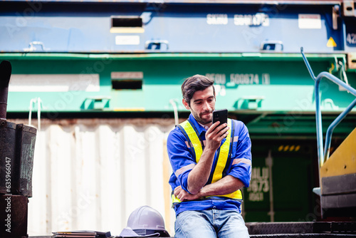 Engineer or Technician texting smartphone cell phone during break time after long hours of working with cargo container yard or engineering site.