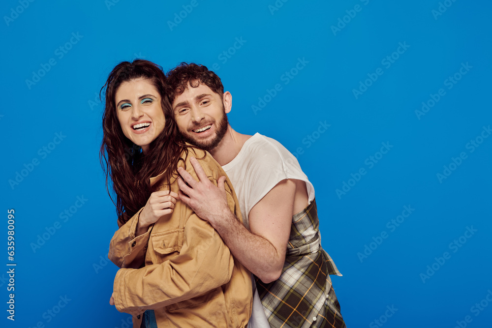 Fototapeta premium cheerful couple in casual wear posing on blue backdrop, bearded man hugging woman, bold makeup