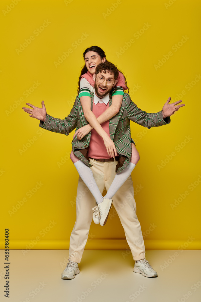 Obraz premium excited couple, funny, young man piggybacking brunette woman on yellow backdrop, student outfits