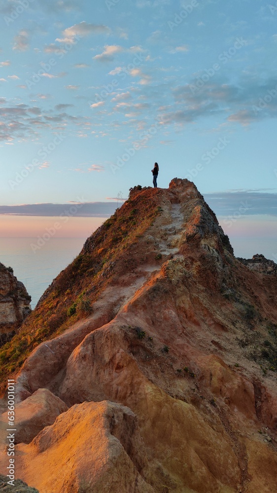 Fototapeta premium Vertical of a woman standing on a rocky mountaintop at sunset at Portugese coastline