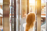 Rear view of blonde girl kid finger pressing button to call elevator in modern residential building. Human child hand pushing buttons of glass lift call, sunshine. Up to high floor. Copy ad text space