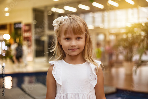 Close-up shot of adorable blonde little girl 5-6 year old wearing in white dress, looking at camera. Portrait of charming smiling girl kid posing indoors. Happy childhood concept. Copy ad text space