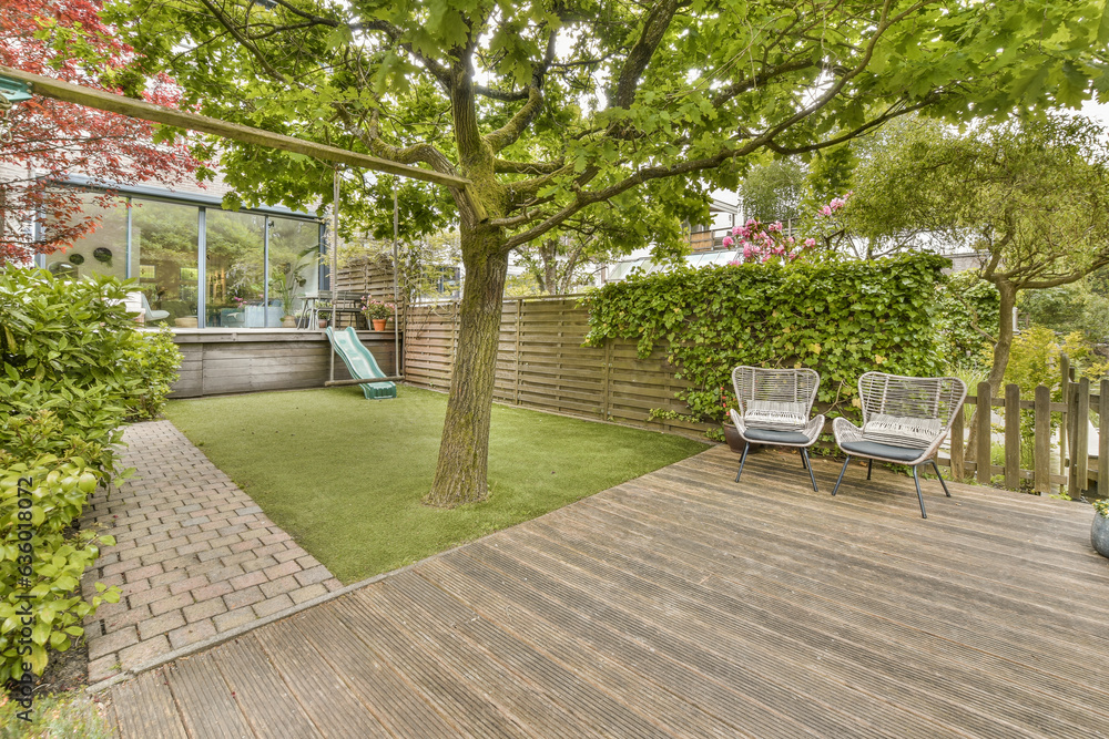 a backyard with wooden decking and green plants on the side of the house there is a tree in the yard