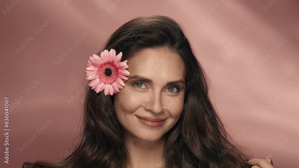 Portrait of a woman with a gerbera flower in her hair in the studio on a pink background. Cosmetic hair care products with extract, flower fragrance. The concept of beauty, cosmetology.