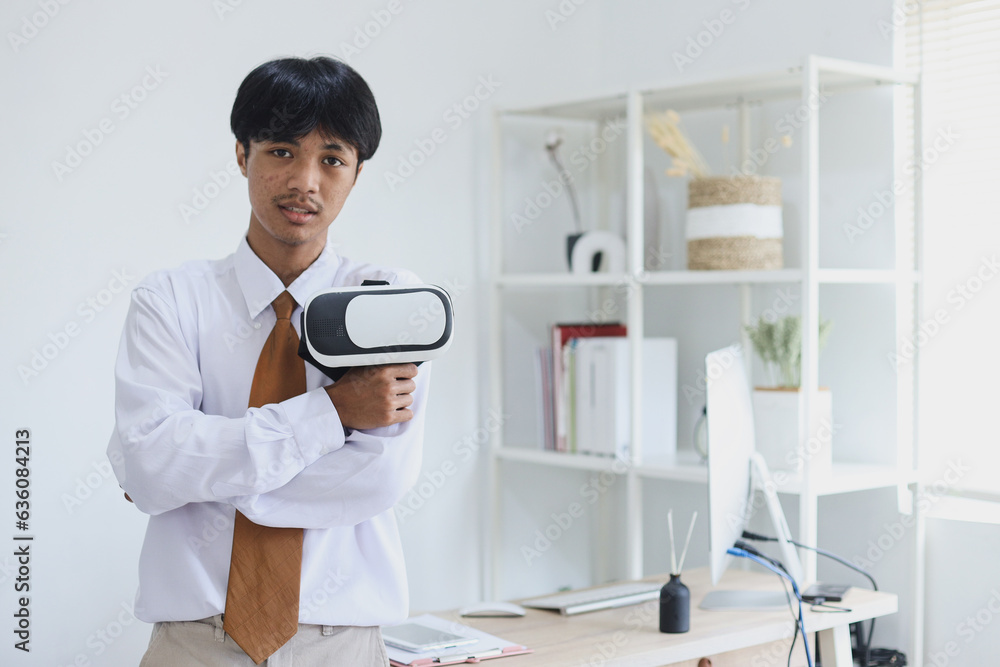 Portrait of young Asian businessman standing in modern office and holding vr glasses while looking at camera confidently