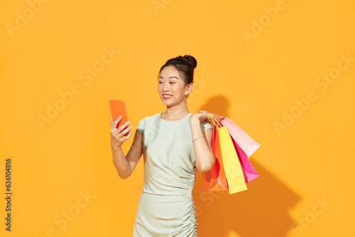 happy young woman holding shopping bags and mobile phone over yellow photo