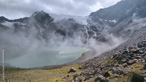 Clouds and fog rolling over remote high alpine lake with huge glacier above and sunlight breaking through photo