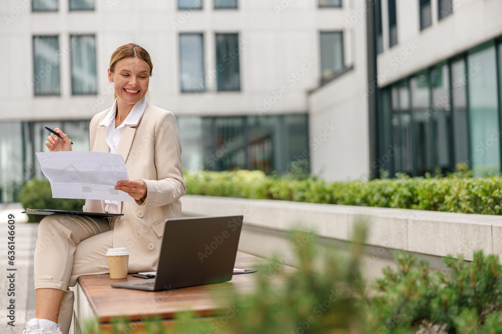 Businesss woman working with documents and use laptop sitting outside on modern building background Businesss woman working with documents and use laptop sitting outside on modern building background