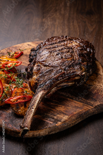 A large fried piece of meat on the bone Tomahawk lies on a wooden board with grilled vegetables