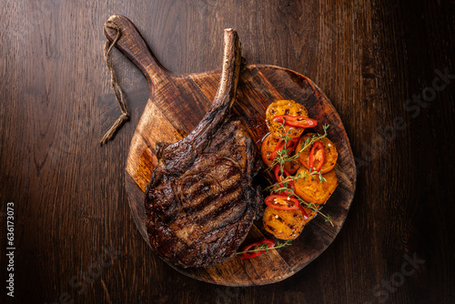 A large fried piece of meat on the bone Tomahawk lies on a wooden board with grilled vegetables