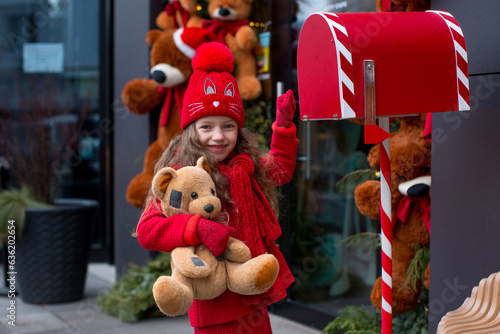A child girl dressed in red stands next to a mailbox and holds a teddy bear in her hands. Merry Christmas