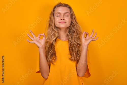 Young attractive serene Caucasian woman in casual t-shirt closes eyes and makes meditative yoga gestures to relax and sort out thoughts after doing difficult work stands on plain orange background.