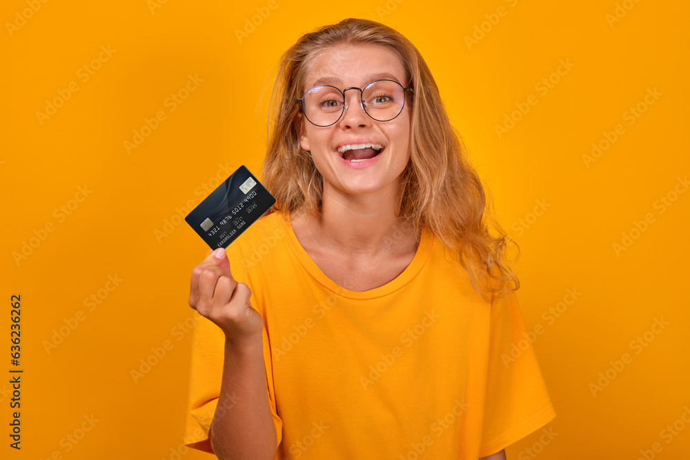 Young cheerful pretty Caucasian woman showing credit card and laughing recommending use of cashless payment to make purchases or get profitable loan from bank stands on orange studio background. Young cheerful pretty Caucasian woman showing credit card and laughing recommending use of cashless payment to make purchases or get profitable loan from bank stands on orange studio background.