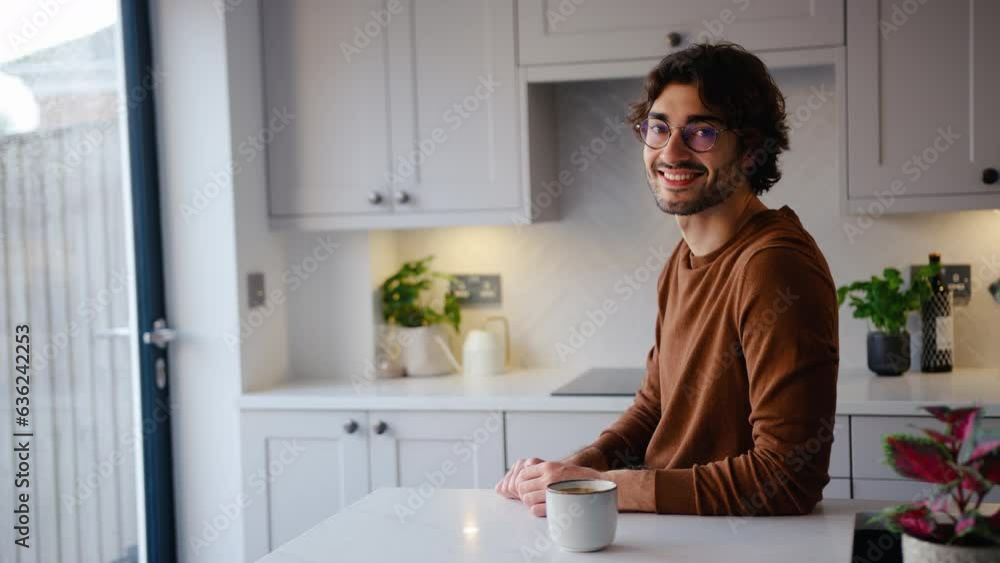 Portrait of smiling young man wearing glasses taking a break with cup of coffee sitting at kitchen counter at home with copy space on left side of frame - shot in slow motion Portrait of smiling young man wearing glasses taking a break with cup of coffee sitting at kitchen counter at home with copy space on left side of frame - shot in slow motion