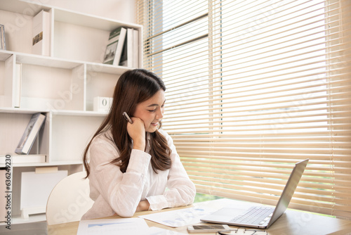 Woman using laptop to work or do homework at home with smiling face in her office, Creating happiness at work with a smile, Live performance or vdo call with laptop, Work from home. photo