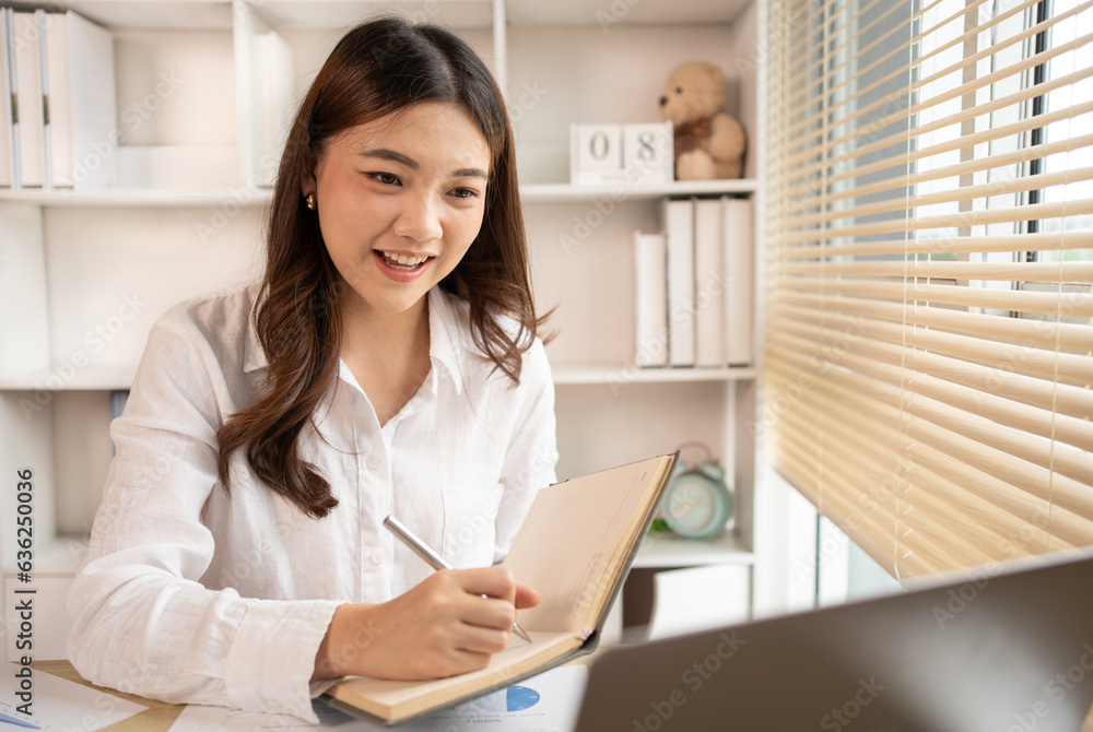 Beautiful young asian woman watching live video or video call of teacher teaching on laptop in her home, Take notes of important conversations and messages during the teacher's teaching. Beautiful young asian woman watching live video or video call of teacher teaching on laptop in her home, Take notes of important conversations and messages during the teacher's teaching.