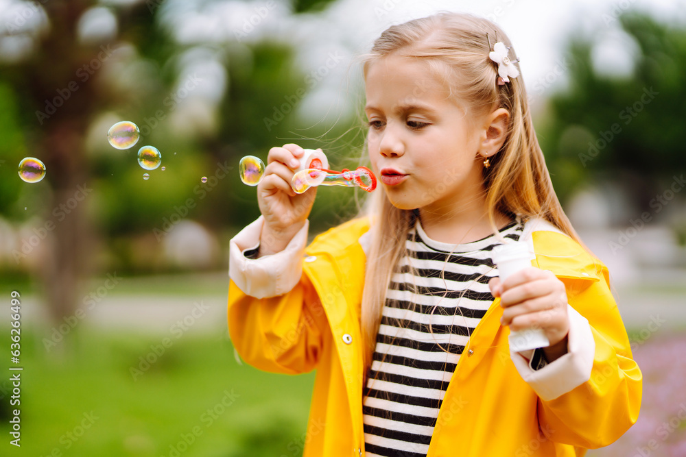 Cheerful little girl in a yellow raincoat blows soap bubbles, plays on a walk in the park in autumn. Childhood, outdoor activities, weekends. Cheerful little girl in a yellow raincoat blows soap bubbles, plays on a walk in the park in autumn. Childhood, outdoor activities, weekends.