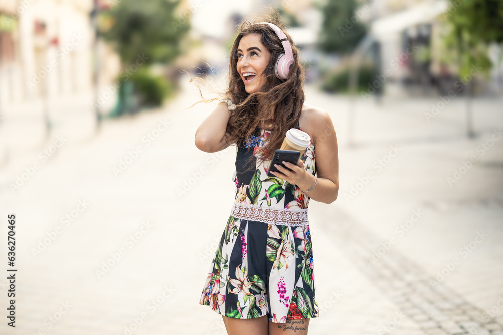 Young beautiful smiling female tourist is using headphones listening to music and holding takeaway coffee in her hand