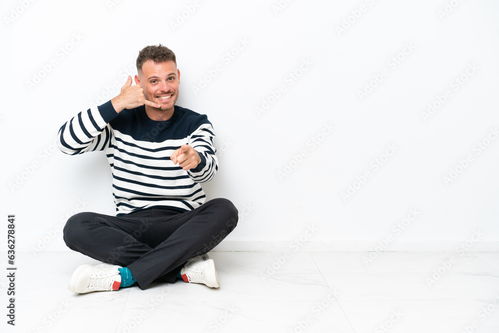 Young man sitting on the floor isolated on white background making phone gesture and pointing front Young man sitting on the floor isolated on white background making phone gesture and pointing front