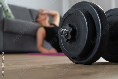 Tired woman resting on a floor after heavy intense exercise. Focus on gym dumbbell barbell equipment. House fitness workout, sport training concept. Exhausted sporty female taking a break at home.