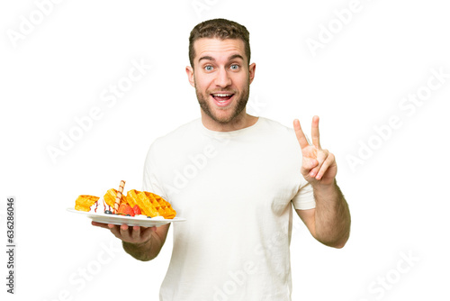 Young handsome blonde man holding waffles over isolated chroma key background smiling and showing victory sign