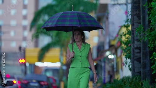 Pensive woman walking in the rain holding umbrella, strolling in urban environment sidewalk during bad weather. Person walks forward in street during rainy afternoon