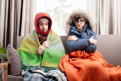 Tablou pe pânză Sad middle age couple in warm clothes getting cold on the sofa in their living r