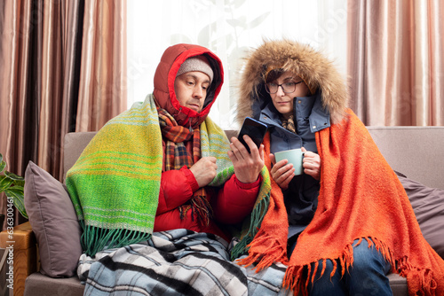 Middle age family in parkas drinking coffee on the sofa while using smartphone in their cold living room