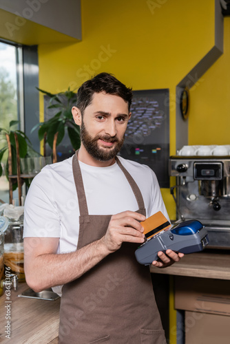 smiling barista using payment terminal and credit card while looking at camera in coffee shop