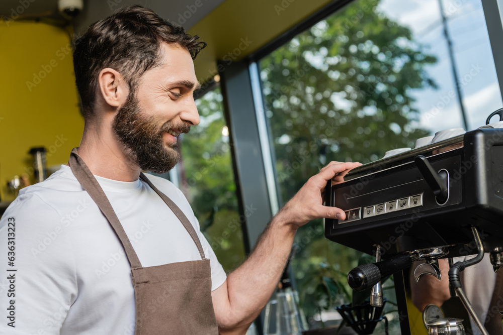 side view of smiling and bearded barista in apron using coffee machine while working in cafe