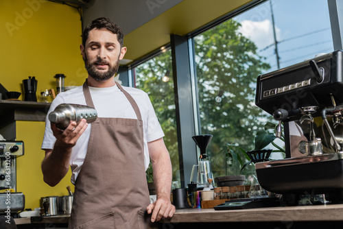 bearded barista in apron using shaker while working near coffee machine in coffee shop